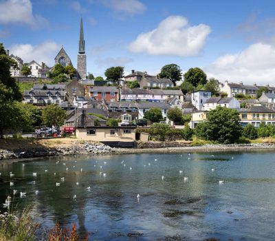 A scenic view of a quaint village with a prominent church spire, nestled along a waterfront dotted with birds.